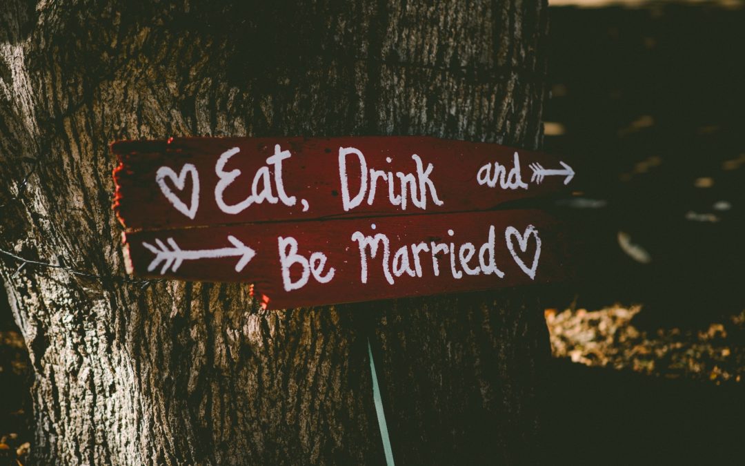 A red sign leaning against a tree that reads “Eat, Drink, and Be Married” in eclectic handwriting.